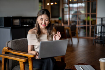 Fototapeta premium Asian woman raising a hand on video call via laptop while sitting on armchair in cafe or coffee shop.=