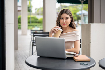 Long hair asian woman looking into camera while holding a coffee cup with laptop aside on cafe table