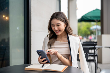 Asian woman smiling at smartphone while sitting with a book on black table in a cafe or coffee shop.