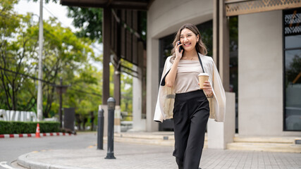 Asian woman smiling during a phone call walking on sidewalk in front of building with coffee in hand