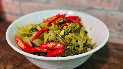Delicious stir-fried Hakka pickled mustard greens with minced pork, garlic, red chili — rustic red brick and wooden table background at a Hakka restaurant in Miaoli countryside, Taiwan