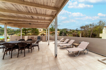 Spacious sunlit terrace featuring sun loungers and wooden dining table under pergola, surrounded by green hills, ideal for relaxing or entertaining outdoors.
