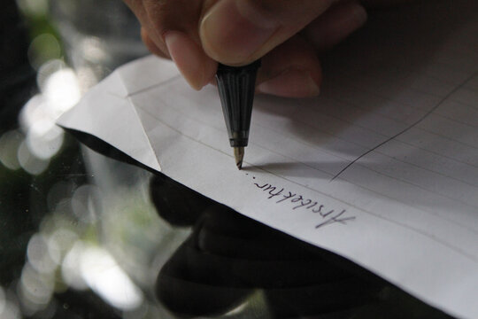 A close-up of a hand writing on a piece of paper. A person's hand holds a black pen and actively writes on white paper, where some handwriting is already present. The background is softly blurred