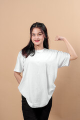 Fototapeta premium Smiling young Asian woman in a white t-shirt and black jeans flexing her biceps in front of a beige background, symbolizing female empowerment, confidence, and positive energy.