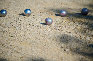 Close-up of metal petanque balls on a gravel playing field during a sunny day. Some balls have distinctive pink dot patterns. Game in progress with shadows on the ground