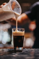 Freshly brewed dark coffee being topped with creamy milk foam by a barista on a marble counter, showcasing the artful process of coffee crafting in a cozy cafe environment.