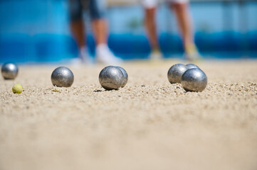 Petanque balls and small yellow jack scattered on sandy court during an outdoor game, with blurred players in the background. Focus on precision and friendly competition