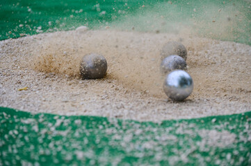 Petanque precision shooting competition moment with metal balls hitting target in sand, dust flying on impact. Focus on accuracy and dynamic play
