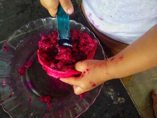 A child's hand is seen scooping bright red dragon fruit from its skin into a glass plate, creating...