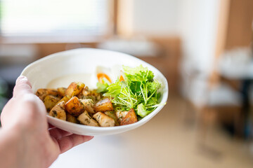 A hand holds a white bowl with grilled chicken, roasted potatoes, fresh green salad, and a halved...
