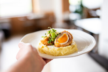 A hand holds a white plate with mashed potatoes topped with a breaded, fried egg wrapped in meat, garnished with microgreens. The background is blurred, suggesting a restaurant setting.