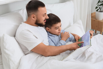 Happy father with his son in pajamas reading book on bed at home