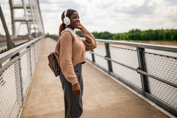 Happy young black woman is walking on bridge foot path, listening music and smiling. She is...