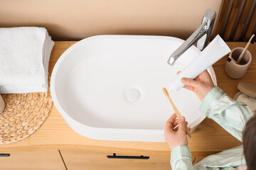 Young woman applying toothpaste on brush near sink in bathroom, top view