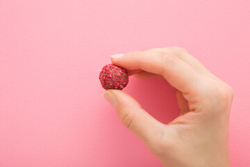 Young adult woman hand fingers holding dark chocolate truffle with pink nut sprinkles on light pink background. Pastel color. Sweet snack. Closeup. Front view.