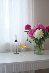 A chic vase of vibrant pink peonies, a refreshing wine drink, copybook with pen, and an elegant candle holder in a serene indoor setting