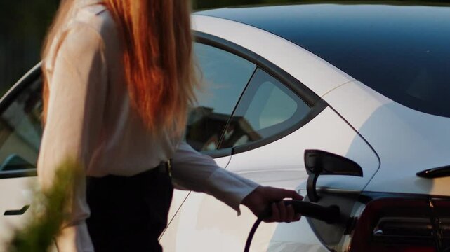 Young woman charges electric car at station during evening hours in a green environment