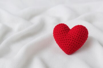 A crocheted red heart rests on a soft white fabric, symbolizing love and warmth with a handmade touch