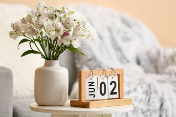 Wooden calendar with date 2 October and vase of beautiful flowers on table near sofa in living room