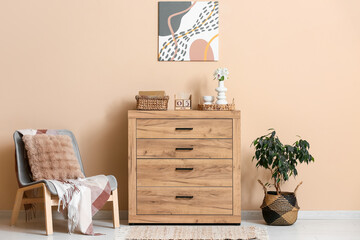 Interior of living room with armchair, wooden cube calendar, vase of flowers and home decor on commode near beige wall