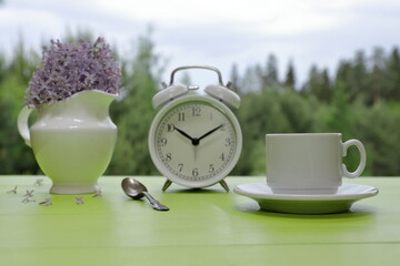 A white cup of coffee stands next to a retro alarm clock and a white jug of lilac flowers on a green wooden desk against a summer woodland background. 