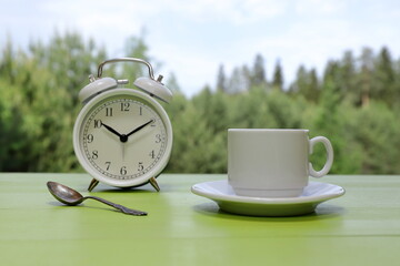 A white cup of coffee stands next to a retro alarm clock on a green wooden desk against a summer woodland background.