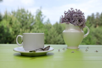 A white cup of coffee stands next to a white jug of lilac flowers on a green wooden desk against a summer woodland background.