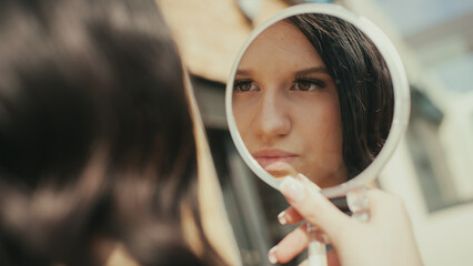 A young woman looks into a handheld mirror while applying makeup. She is fashionable and carefully poses for an upcoming social media post