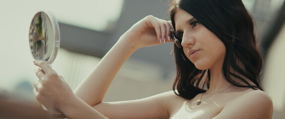 A young woman looks at herself in a hand mirror. She poses, perhaps for content on social media, in a modern, urban environment. She seems fashionable and modern