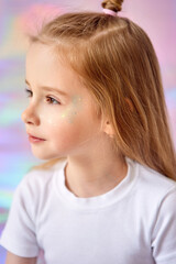 A close-up portrait of a charming little girl with long blond hair. She looks thoughtfully into the distance. The model's face is decorated with glitter. The background has gentle pastel shades.