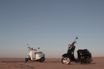 Real photo of two retro scooters parked in the golden sands of the Namib Desert at sunset. A warm,...