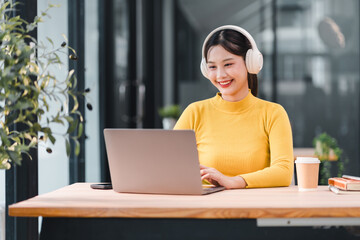 Young woman in yellow sweater smiling while working on laptop with headphones, sitting at modern office desk with coffee cup, books, and greenery, feeling positive and focused