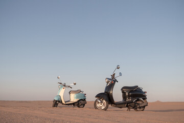 Real photo of two retro scooters parked in the golden sands of the Namib Desert at sunset. A warm,...