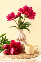Vase with beautiful peony flowers and cup of hot coffee on white wooden against yellow background