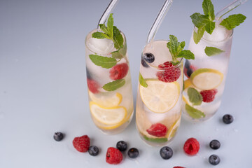 refreshing cocktail trio with mint, lemon, and berries in glasses topped with ice and glass straws on light surface. sparkling, elegant, thirstquenching, zesty, freshness, bright, colorful