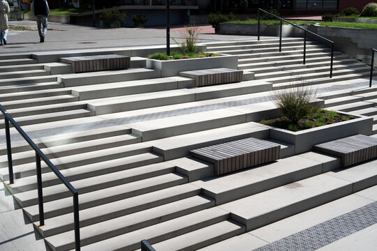 sunlit urban plaza staircase composed of staggered concrete terraces, integrated wooden benches, green planters, and sleek black handrails, creating a minimalist geometric landscape for pedestrians
