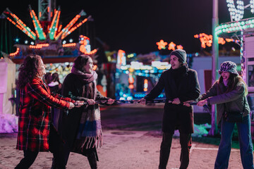 A group of friends playfully pulling a rope in a vibrant winter carnival at night, surrounded by...