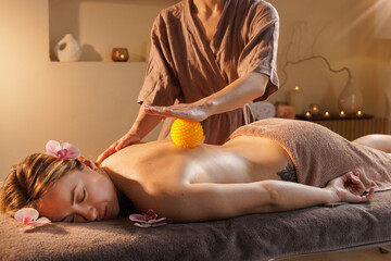 Spa therapist performs trigger point massage using a spiky massage ball on woman's back to relieve...