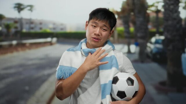 Young man holding soccer ball on city street, wearing a blue scarf, exhibits outdoor fan enthusiasm, showcasing sports passion and joyful expression.