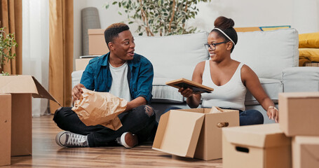 A husband and wife unpack boxes in their flat, sitting close on the floor. They reveal a framed...
