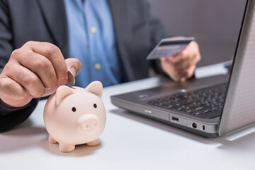 A person saves money in a piggy bank while holding a credit card beside a laptop, symbolizing...