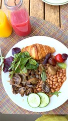 Beans and mushrooms fill half plate with croissant, cucumbers, cherry tomato, grapes, olives, red lettuce and bright green leaves, glass bottles of juice behind on wood table. Vegan balance.
