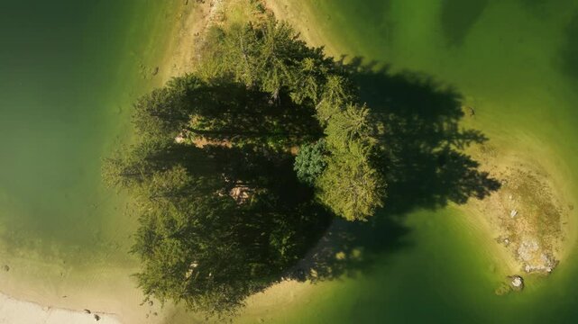 Aerial view captures a small island surrounded by tranquil Lake Predil waters, featuring dense greenery and trees with soft sandy borders. The peaceful scene invites relaxation and exploration.