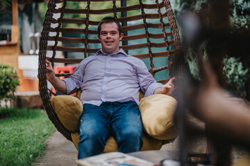 A young man with Down syndrome is sitting comfortably in a swing chair outdoors, smiling and...