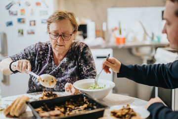 An elderly woman serves food during a meal with a younger companion, capturing themes of family...