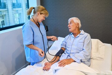 Obraz premium Nurse taking blood pressure of elderly man in hospital bed