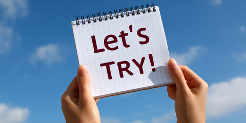 Hands hold notepad against blue sky, displaying "Let's Try!" message, conveying encouragement and motivation for new beginnings or challenges