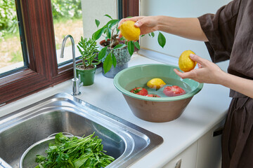 A woman washes fresh lemons, tomatoes, and leafy greens in a kitchen sink, preparing healthy ingredients near a sunlit window.