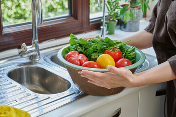 A woman washes fresh lemons, tomatoes, and leafy greens in a kitchen sink, preparing healthy ingredients near a sunlit window.