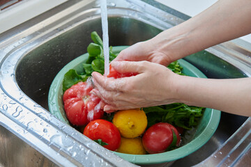 A woman washes fresh lemons, tomatoes, and leafy greens in a kitchen sink, preparing healthy ingredients near a sunlit window.